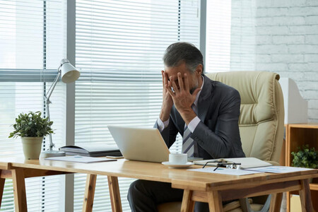 Hombre de negocios en un despacho, frente a un portátil, cubriéndose el rostro con las manos por un error